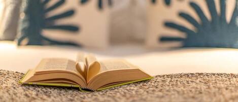 Un livre ouvert, posé sur une surface texturée, vraisemblablement un lit ou un canapé, avec un arrière-plan flou où l’on aperçoit un coussin orné d’un motif de feuilles.