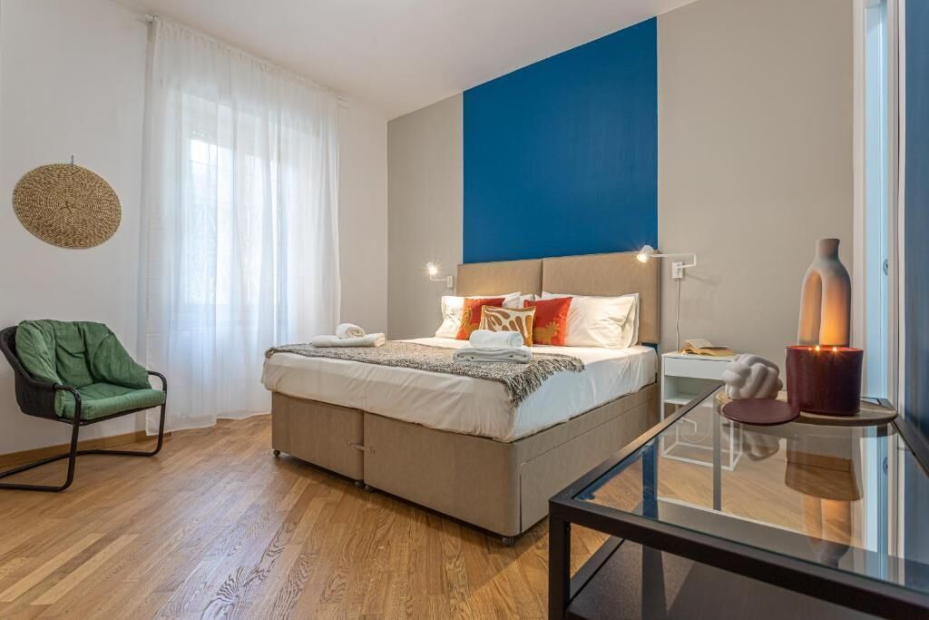 Un dormitorio con cama, una silla, una mesita de noche, una lámpara y una ventana con cortinas.