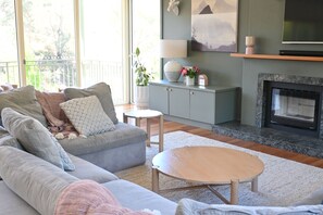 A living room with a grey sofa, a round coffee table, a fireplace, and a view of greenery outside. - The River House - Merrijig (Merrijig)