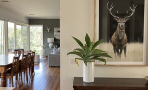 A dining area with a wooden table and chairs, a large window with a view, and a framed deer painting on the wall. - The River House - Merrijig (Merrijig)