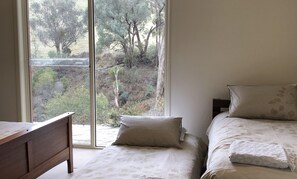 A bedroom with a bed, a nightstand, and a window with a view of trees and a hillside. - The River House - Merrijig (Merrijig)