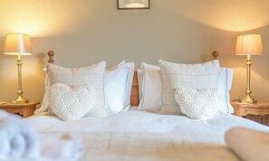 Cama bem arrumada com almofadas em formato de coração, duas luminárias de cabeceira e um objeto emoldurado na parede.