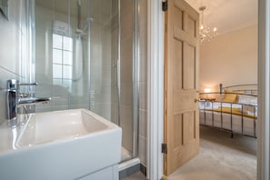 Une salle de bain moderne avec une douche à l’italienne en verre, un lavabo blanc et une porte en bois donnant sur une chambre à coucher équipée d’un lit et d’une table de chevet.