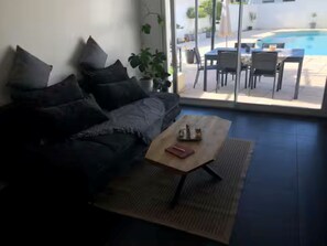 A living room with a sofa, a coffee table, and a view of an outdoor pool area. - Maison Vallée: Swimming Pool / Groupama Stadium / Euroexpo / LDLC ARENA / Access to Downtown (Décines-Charpieu)