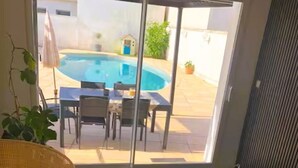 A room with a sliding glass door leading to a pool area, a sofa, a table with a book and a remote control, and a wicker lamp. - Maison Vallée: Swimming Pool / Groupama Stadium / Euroexpo / LDLC ARENA / Access to Downtown (Décines-Charpieu)