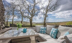 Uma área de estar externa com mesa de vime, almofadas e vista para o campo.