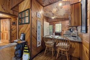 Interior de una cabaña de madera con zona de cocina, dos sillas de madera, un ventilador de techo y un letrero que dice 'HIVE'.