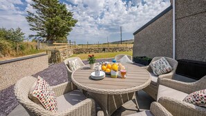 A round outdoor dining table with wicker chairs, a stone wall, and a view of the countryside.