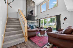 A modern living room with a fireplace, a staircase, and a large window offering a view of the outdoors. - Fairway Getaway Near Mille Lacs 3 Bedroom Home (Onamia)