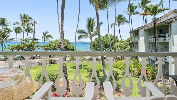 A balcony with white chairs and a table, overlooking a lawn with palm trees and a view of the ocean. - Big Island Kona Islander 245 Condo (Kailua-Kona)