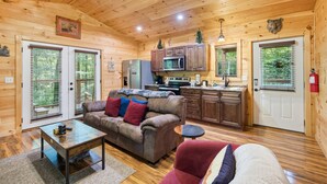Interior de una cabaña de madera con cocina, sala de estar con sofá y mesa de centro, y una puerta decorada con una cabeza de oso.