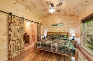 Un dormitorio de cabaña de madera con una cama, dos mesitas de noche, un ventilador de techo y una ventana.