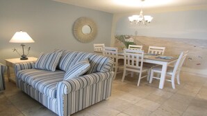 Una sala de estar con un sofá a rayas, una mesa de comedor con sillas, una araña y una pared con un espejo circular y una escena de playa.