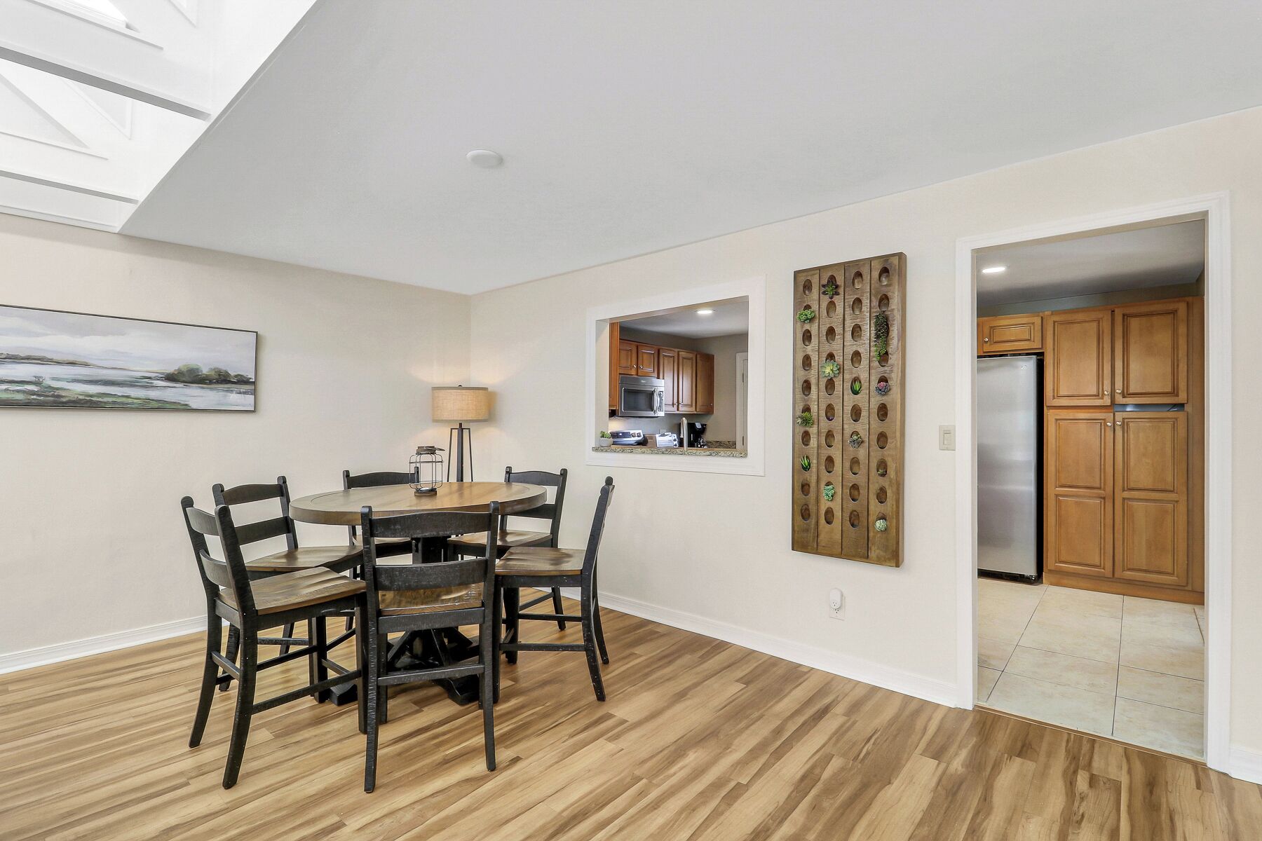 A dining area with a round table and chairs, a kitchen visible through an open door, and a wall-mounted decorative panel.
