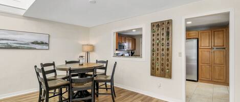 A dining area with a round table and chairs, a kitchen visible through an open door, and a wall-mounted decorative panel.
