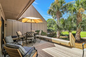An outdoor patio area with a covered seating arrangement, including chairs and a table, and a large umbrella providing shade. - Sunset Rentals | Queens Grant 671 | Dog Friendly (Hilton Head Island)