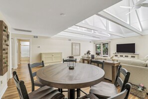 A modern living room with a round dining table, comfortable seating, a flat-screen TV, and a large window providing natural light. - Sunset Rentals | Queens Grant 671 | Dog Friendly (Hilton Head Island)