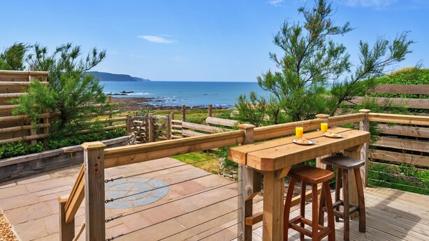 Een houten terras met een tafel en krukken, met uitzicht op de kust.