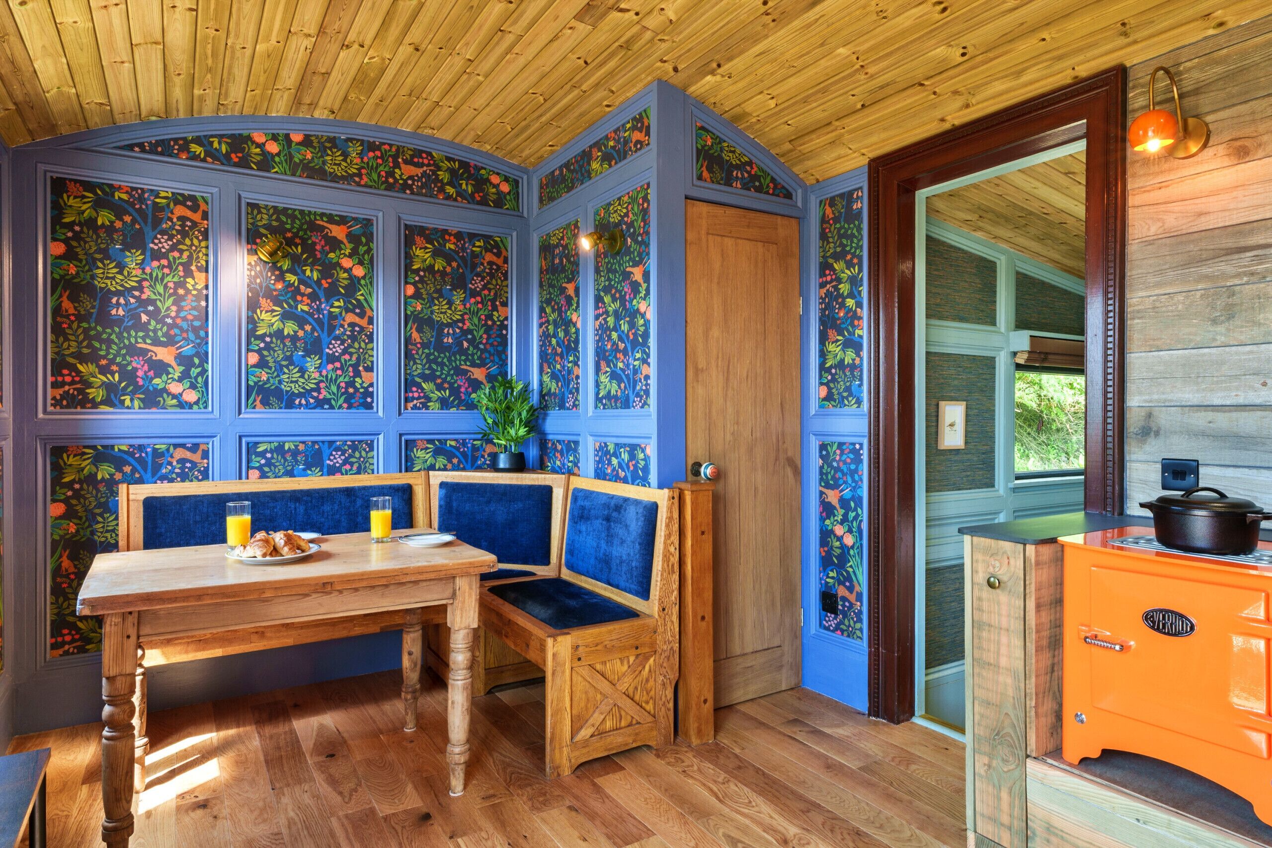 A room with a wooden ceiling, a bench, a table with breakfast items, and an orange stove.