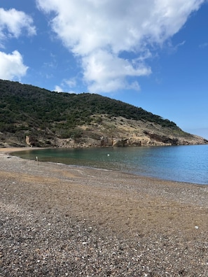 A pebbly beach with a clear view of the sea and a forested hillside in the background.