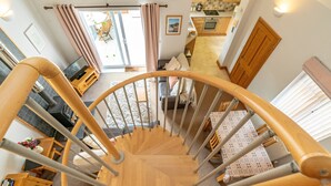 A spiral staircase with a wooden finish, leading to a living area with a sofa, a dining table, and a kitchen in the background. - Bwthyn-Bwlch -  a self catering cottage that sleeps 4 guests in 2 bedrooms (Dinbych)