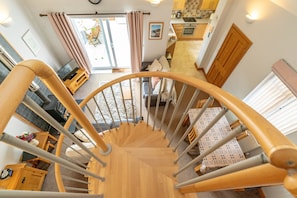 A spiral staircase with a wooden finish, leading to a living area with a sofa, a dining table, and a kitchen in the background.