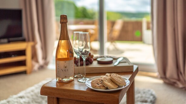 A bottle of wine, two glasses, and a plate of cookies on a wooden table with a view of the outdoors. - Bwthyn-Bwlch -  a self catering cottage that sleeps 4 guests in 2 bedrooms (Dinbych)