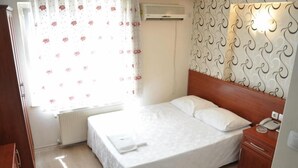 Une chambre d’hôtel avec un lit, une table de chevet avec un téléphone, un radiateur et une fenêtre ornée de rideaux à motifs floraux.