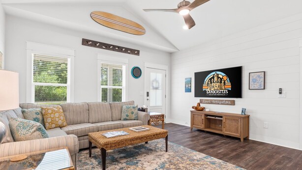 Un salon avec un canapé, une table basse et un téléviseur. Il y a une horloge au mur et une enseigne indiquant « Sandcastles Vacation Rentals ».