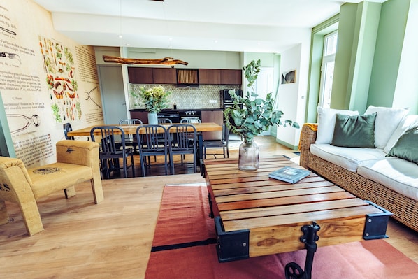 A modern living room with a wooden coffee table, a dining area with chairs, a kitchen in the background, and a green wall.