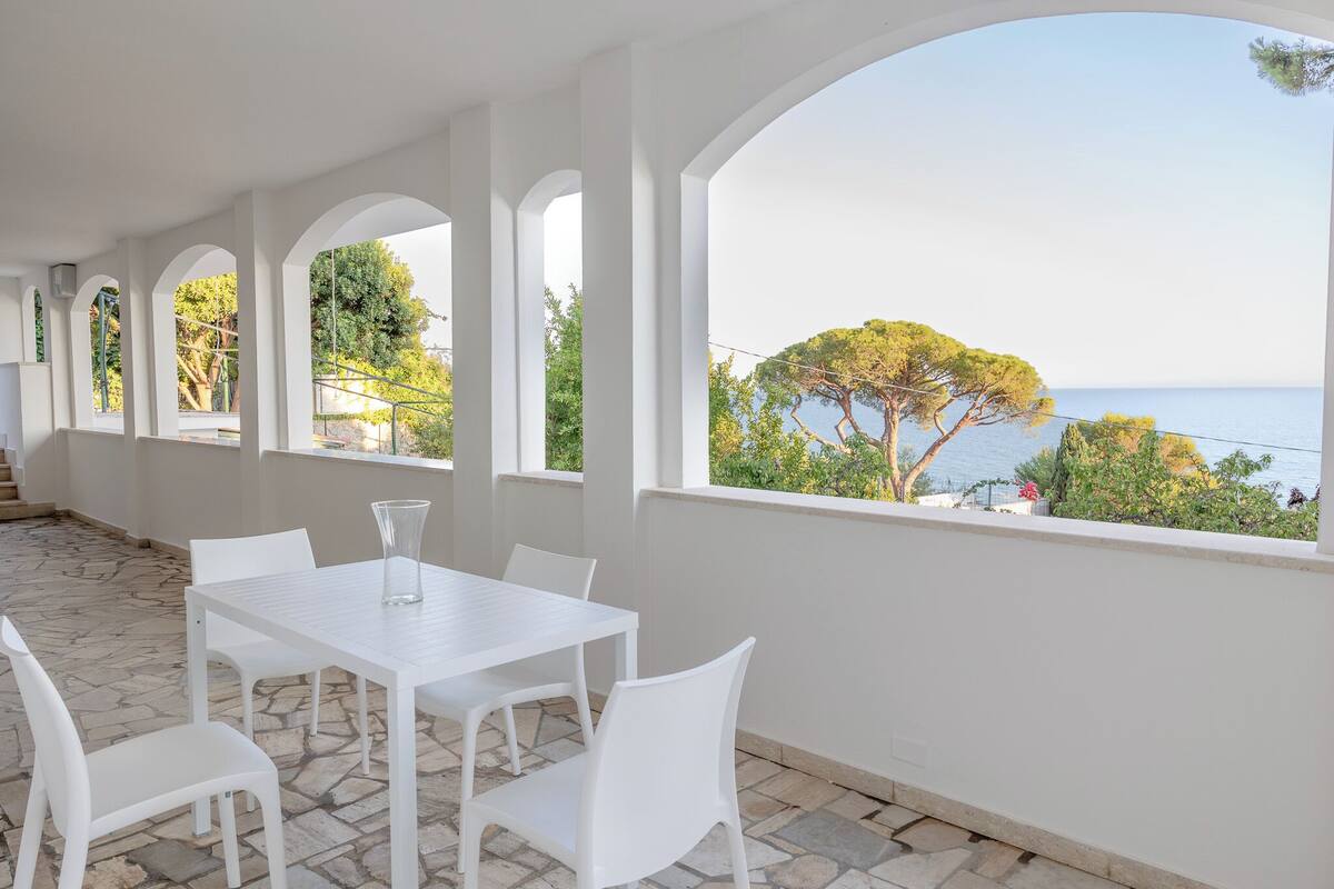 A white dining area with a table and chairs, large windows with a view of the sea and trees, and a stone floor.