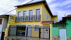 Un bâtiment jaune avec une clôture en métal noir, une enseigne indiquant « PSC Pousada Souza Castro », et un bâtiment vert situé à côté.