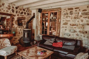 Ein rustikaler Raum mit Steinwand, einem großen Kaminofen, einer Ledersofa, einem Holztisch und einem karierten Sessel.