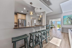 A modern kitchen with bar stools, stainless steel appliances, pendant lighting, and a view to the outdoors.