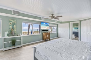 A bedroom with a bed, a ceiling fan, a TV, a dresser, and a view of the beach.