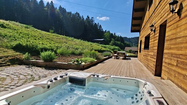 A hot tub with a wooden deck, a wooden building with windows, and a forested hillside in the background.
