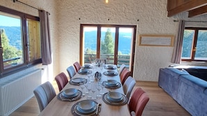 A dining area with a long table set for six, wooden beams on the ceiling, and a view through large windows.