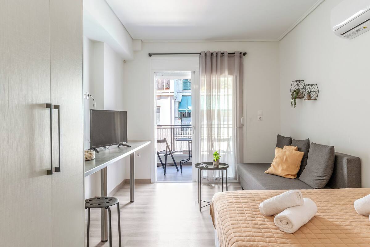 Una habitación de hotel moderna con una cama, un televisor, una mesita con una planta, un balcón con una silla y un sofá con cojines.