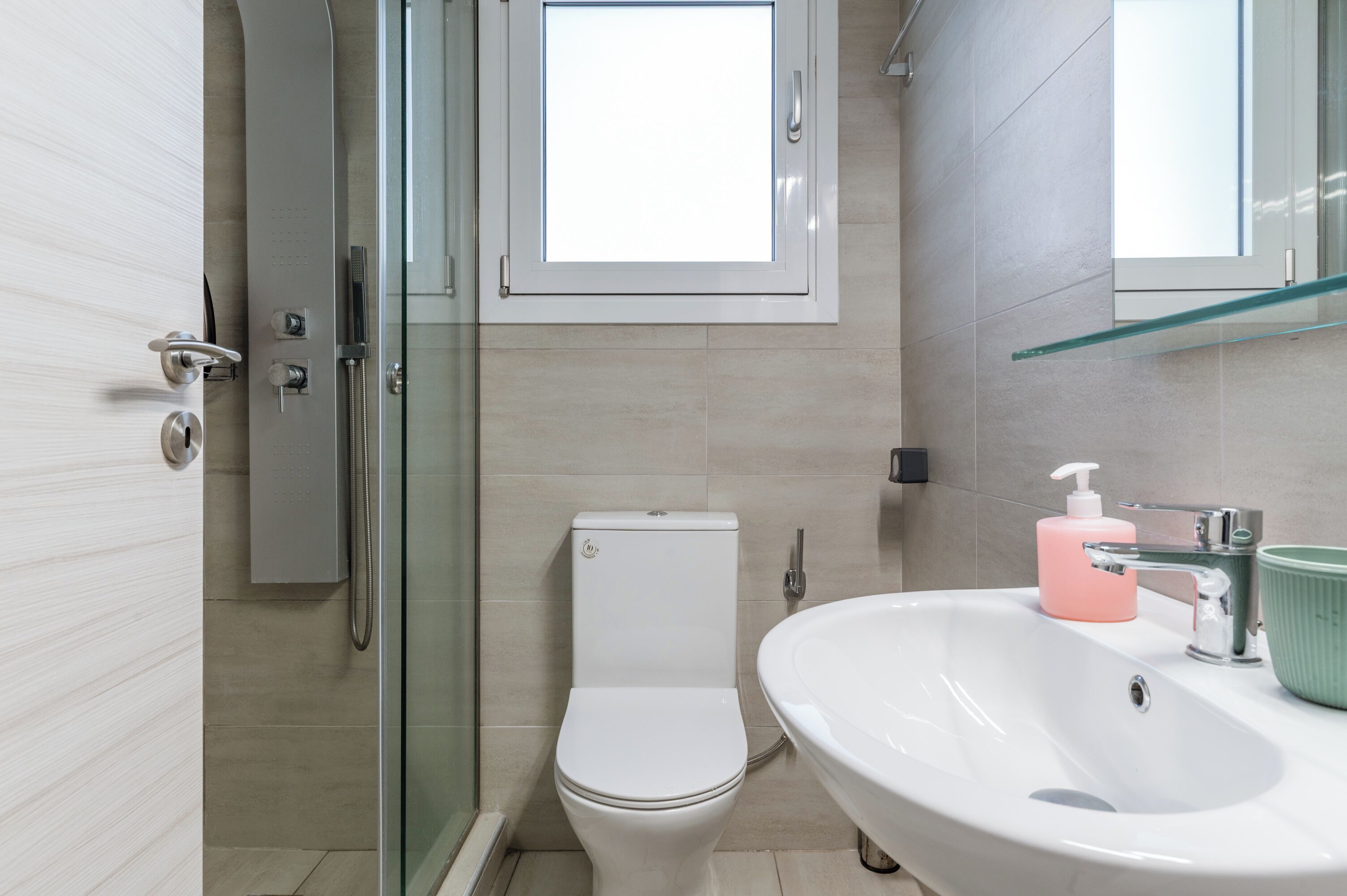 A modern bathroom with a glass shower enclosure, a toilet, a sink with a green basin, and a window above the toilet.