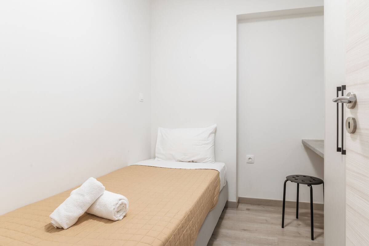 Una habitación pequeña con una cama individual, una colcha color beige, una almohada blanca y una toalla blanca enrollada sobre la cama. Hay un taburete pequeño y una puerta a la derecha.