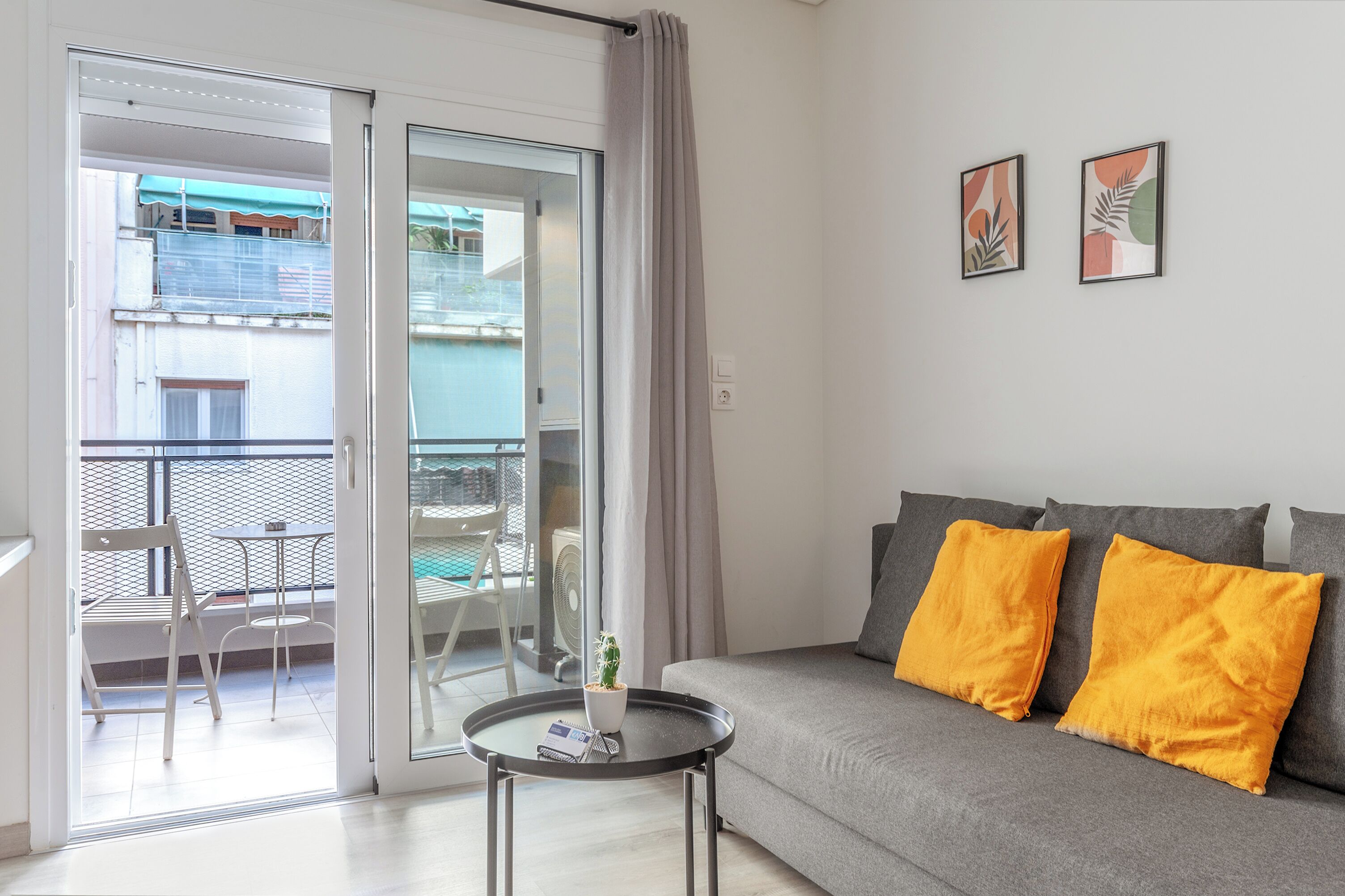 A living room with a grey sofa, two yellow cushions, a small round table with a book and a plant, and a sliding glass door leading to an outdoor balcony.