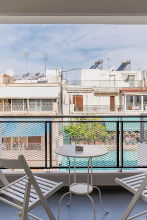 Un balcón con dos sillas blancas y una pequeña mesa redonda, con vista a un patio que tiene una piscina y edificios al fondo.
