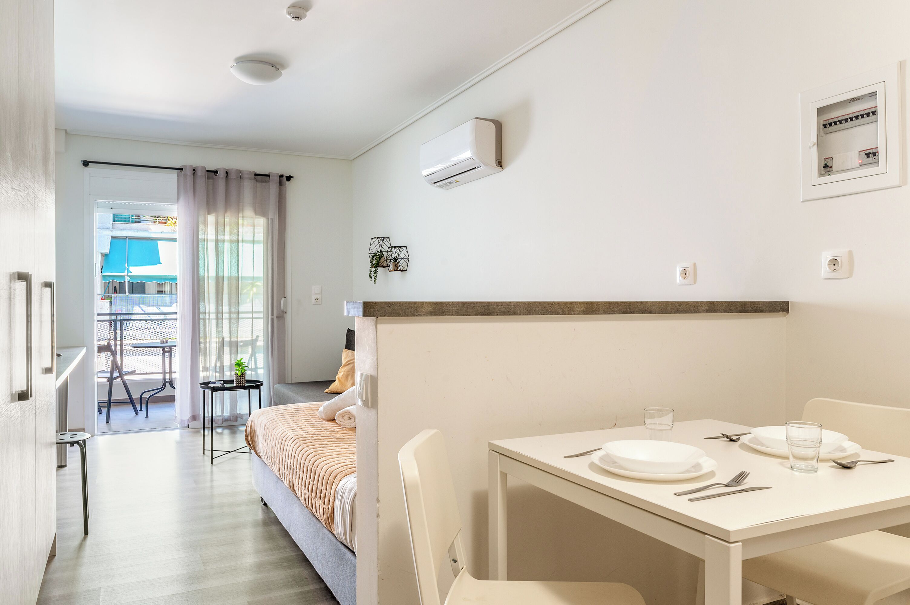 A hotel room with a bed, a dining table set for two, a small table with a plant, and a balcony with a view.