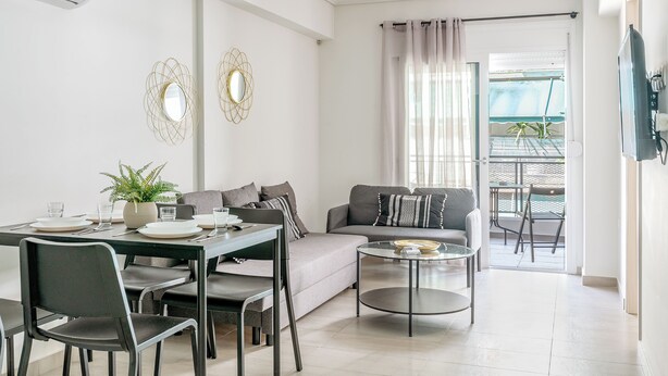 A modern living room with a dining area, a sofa, a coffee table, and a balcony with outdoor furniture. - Elvita Apartments 4 Athens (ATHINA)