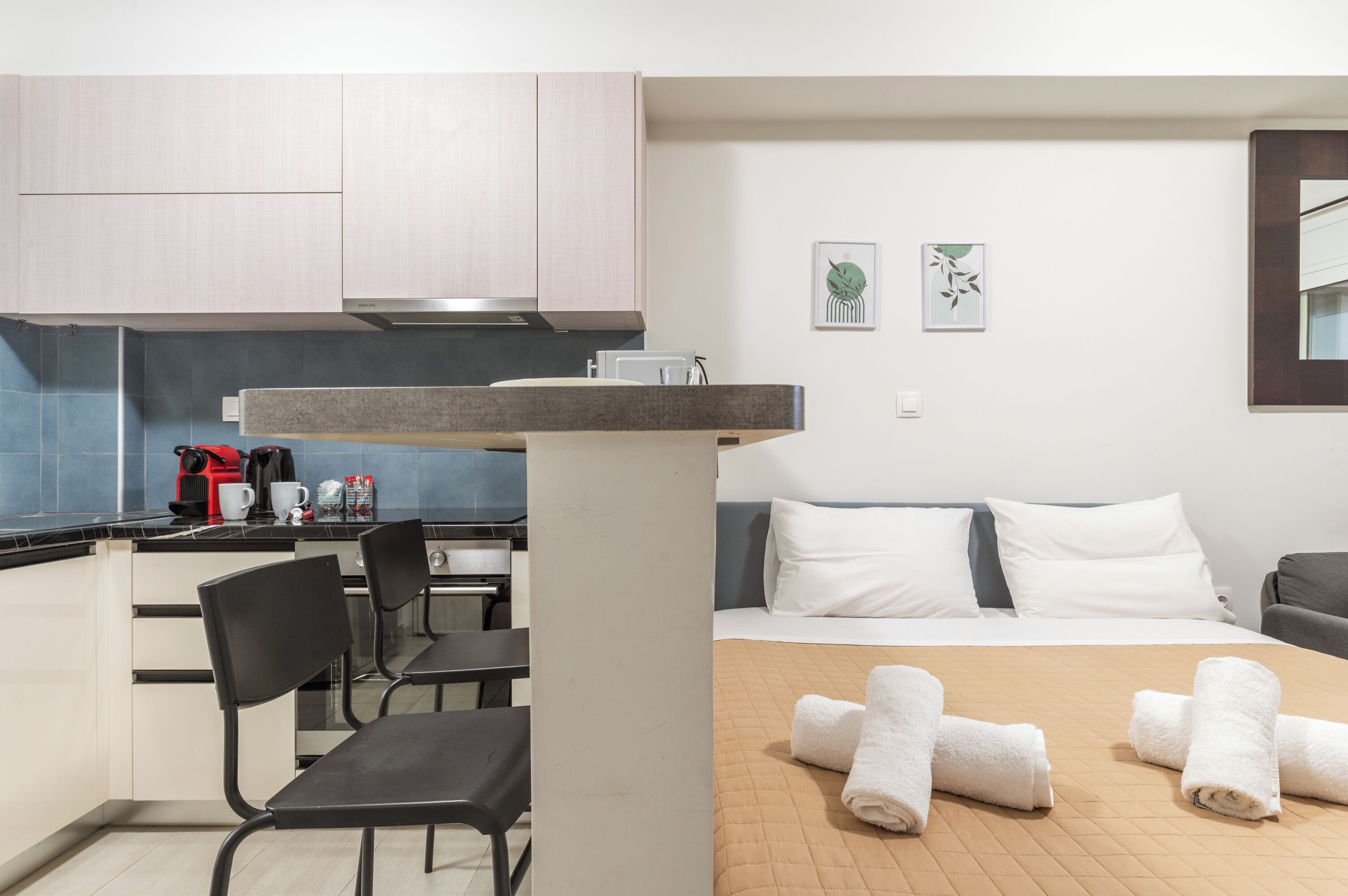 A hotel room with a bed, a desk with a chair, a kitchenette, and two wall art pieces.