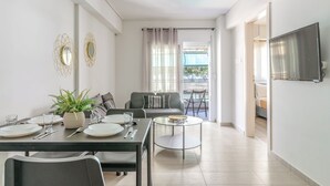 A modern living room with a dining area, a sofa, a TV, and a balcony with curtains. - Elvita Apartments 4 Athens (ATHINA)