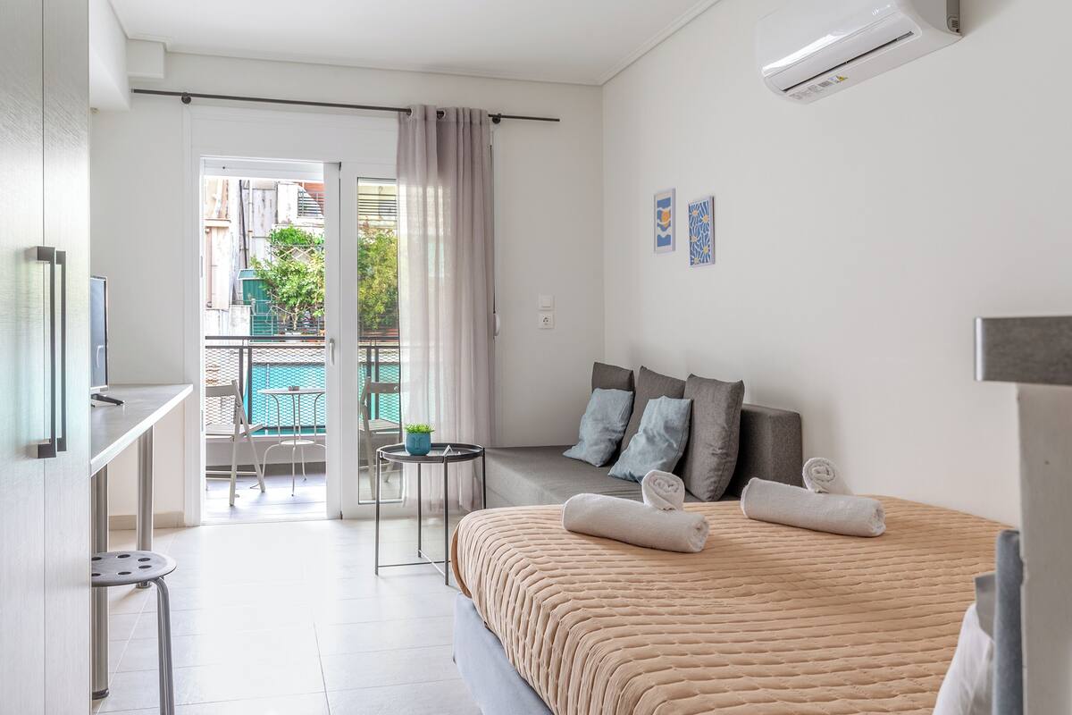 Una habitación de hotel moderna con una cama, una mesita, una silla y un balcón con vistas a edificios y zonas verdes.