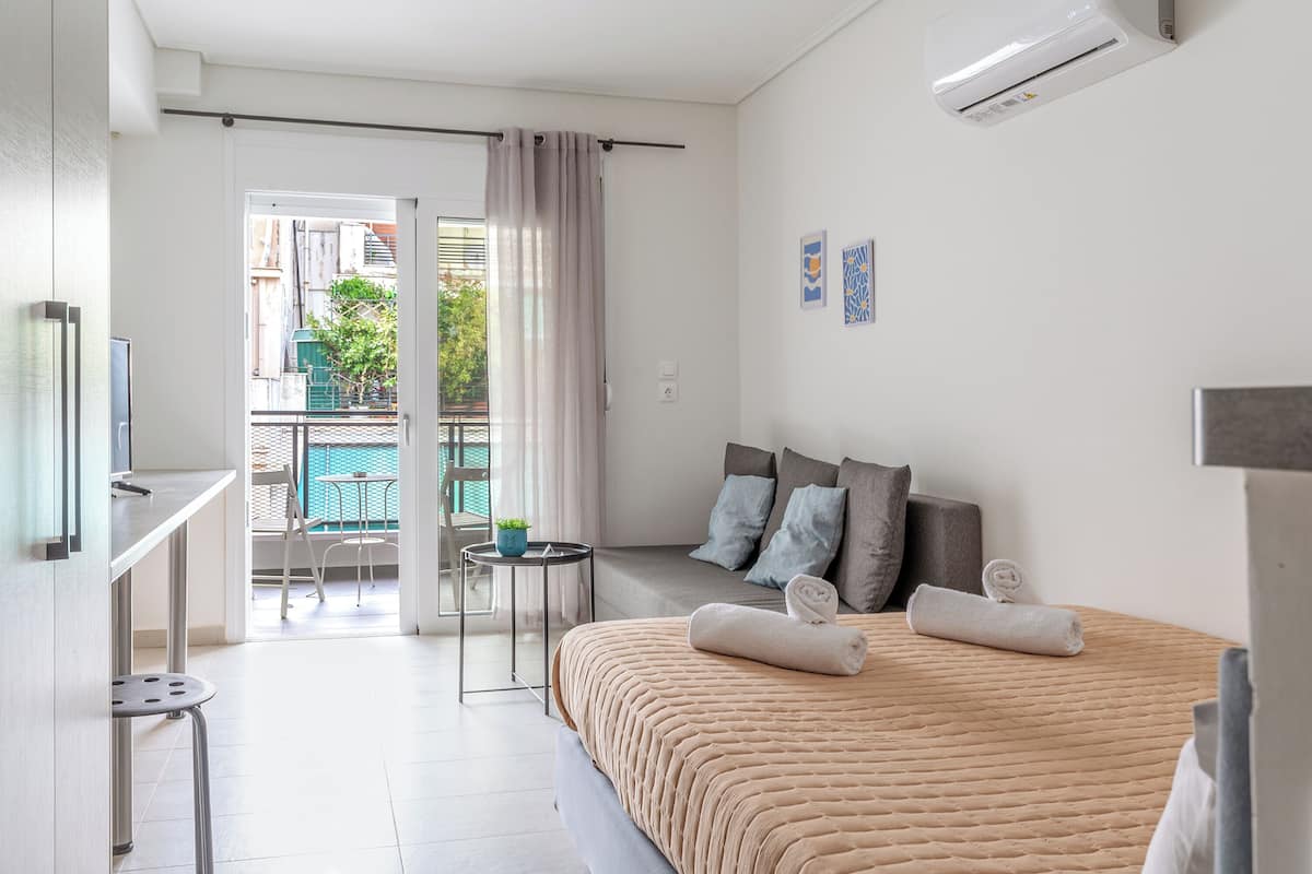 Une chambre dâhĂŽtel moderne, Ă©quipĂ©e dâun lit, dâune petite table, dâune chaise et dâun balcon offrant une vue sur les bĂątiments et la verdure.