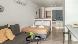 A modern hotel room with a bed, a sofa, a small table with a plant, a kitchenette, and a bathroom visible through an open door. - Elvita Apartments 4 Athens (ATHINA)