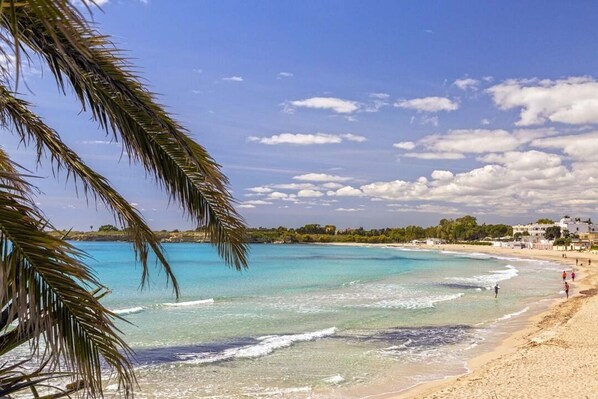 A beach with clear turquoise water, a sandy shore, and a few buildings in the background. - Villa Thalassa, for a perfect stay, 500 meters from the sea (Fontane Bianche)